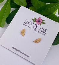 Load image into Gallery viewer, A pair of small gold leaf-shaped stud earrings displayed on a white Lucy &amp; Jane earring card, with green leaves in the background.