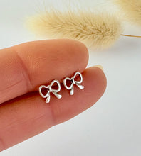 Load image into Gallery viewer, Silver bow-shaped earrings held between fingers with a blurred natural background