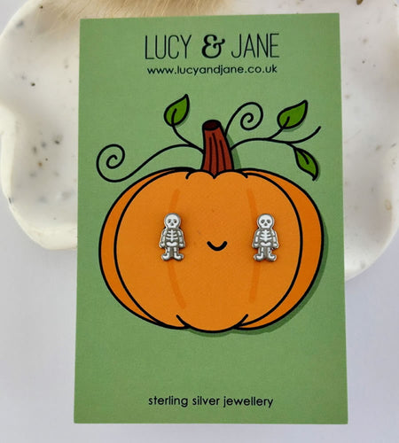Pumpkin-shaped card with skeleton earrings on a green background, featuring the brand 'Lucy & Jane'.