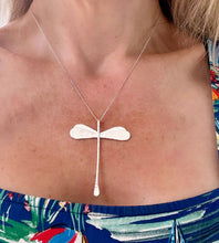 Load image into Gallery viewer, A silver-toned necklace with a dragonfly pendant is worn by a model. The necklace features a delicate chain and a flat, winged cross-shaped pendant with a vertical drop, shown against the model's chest.