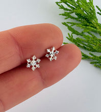 Load image into Gallery viewer, A pair of silver snowflake stud earrings held between two fingers for scale, with green foliage in the background on a white surface.