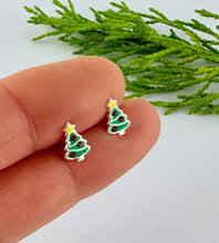 Load image into Gallery viewer, A close-up of a person's fingers holding a pair of small Christmas tree stud earrings with green enamel, silver outlines, and yellow star tops, with green foliage in the background.