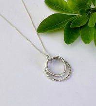 Load image into Gallery viewer, A silver double-ring pendant necklace with one larger open circle and one smaller textured ring, displayed on a white surface next to green leaves.